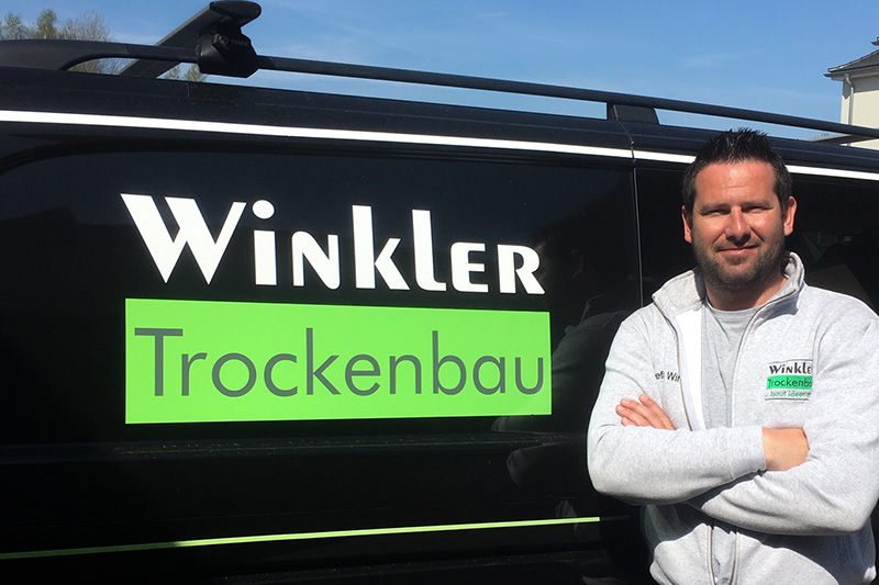 Stefan Winkler, Geschäftsführer von Winkler Trockenbau Meisterbetrieb für Trockenbau & Akustik vor einem schwarzen Firmenfahrzeug mit Logobeschriftung